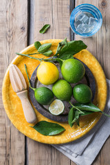limes with lemon on a yellow plate and a wooden table, next to a juicer for citrus and a glass of water