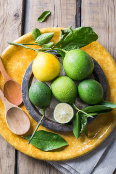 Limes with a lemon on a yellow plate and a wooden table, next to a wooden spoon.
