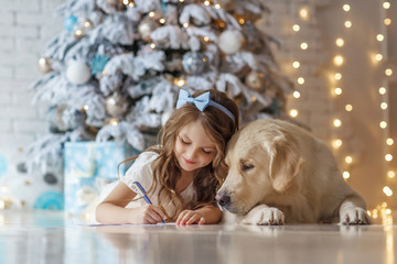 Little cute girl with a golden retriever dog writes letter to Santa near christmas tree