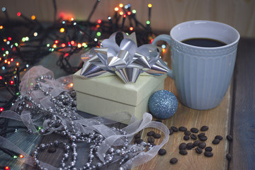 White box with a bow, coffee grains, blue cup of coffee, Christmas tree toy-a blue ball, beads and Christmas lights on a wooden table in a country house