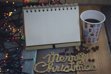 The golden inscription Marry Christmas, a glass and coffee beans, an open notepad for entries and glowing New Year's lights on the table