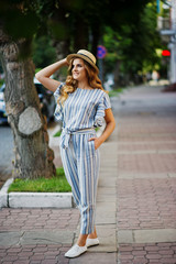 Portrait of a very attractive young woman in striped overall posing with her hat on a pavement in a town with trees in a background.