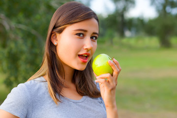 Beautiful teenage girl biting an apple, against green of summer park.