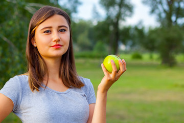 Beautiful teenage girl biting an apple, against green of summer park.