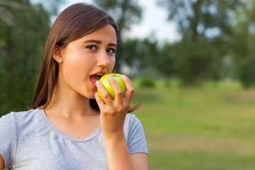 Beautiful teenage girl biting an apple, against green of summer park.