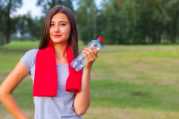 Beautiful teenage girl with a bottle of water, against a green summer park.