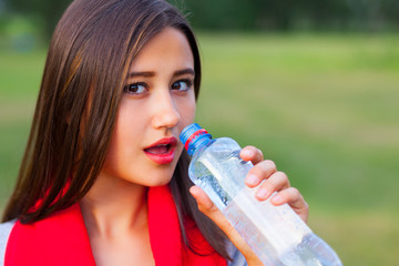 Beautiful teenage girl with a bottle of water, against a green summer park.