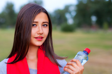 Beautiful teenage girl with a bottle of water, against a green summer park.