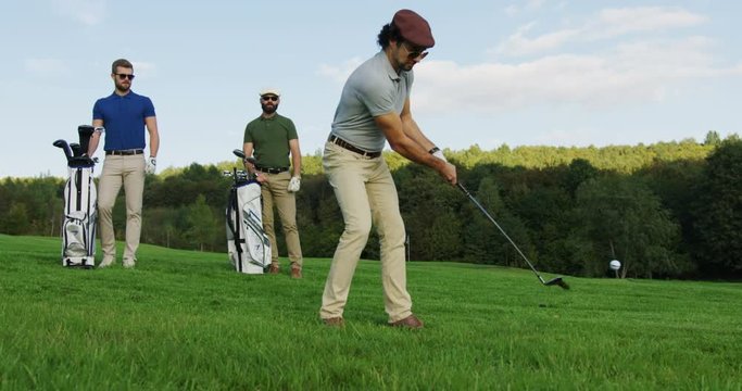 Handsome player in glasses hitting a ball with a club and two men with golf bags watching him on a background. Good sunny weather day. Outdoors