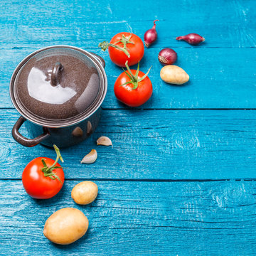 Picture of iron pot, tomato, potatoes, onions on blue wooden background.