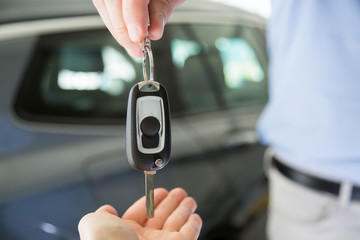 Close photo of male car dealer hand giving a car key to a female person hand. They are standing indoors in a showroom, with cars behind them