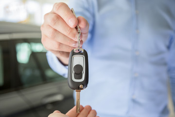 Close photo of male car dealer hand giving a car key to a female person hand. They are standing indoors in a showroom, with cars behind them
