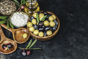 Fresh organic olives placed in wooden bowl with salt, mixed peppers and bottle of virgin olive oil on rustic dark background.Top view with copy space