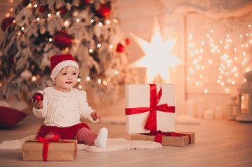 Obraz na płótnie Canvas Cute baby holding Christmas ball wearing Santa Claus hat sitting with presents over Christmas tree in room. Holiday season.