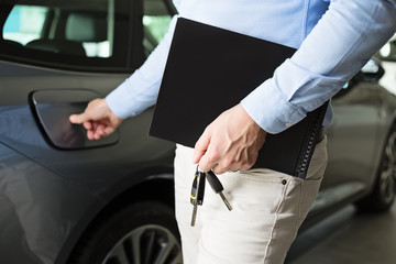 Close up of male adult car dealer standing indoors at car dealership,holding a folder and car keys in his hand, poiting finger towards a fill spout, talking about gas consumption