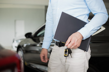 Close up of male adult car dealer standing indoorts at car dealership,holding a folder and car keys in his hand with a new car behind him