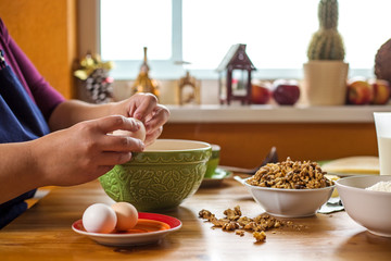 Close up photo of young female hands breaking an egg by the edge of the green bowl, bowl full of wallnuts is placed close by and  ingredients for baking are placed on the table