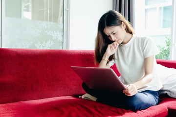 Asian teenage girl using a laptop computer to check her orders online and drinking coffee on a red sofa in her home. She stressed that the sales slump.