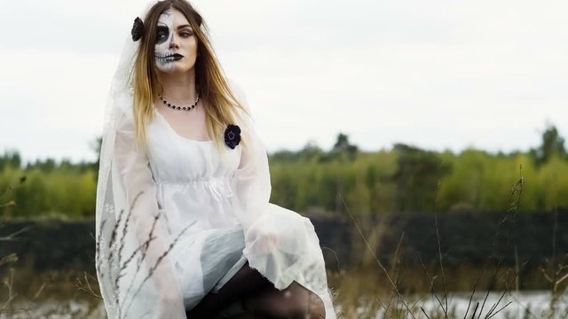 The young woman with frightening make-up of dead bride for Halloween dressed in a white wedding gown sitting in a field of burned grass. 4K