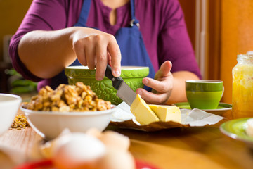Close up photo of young female sitting behind a wooden kitchen table, focus on her hand holding a knife and cutting a slice of butter. Ingredients for baking on the table.