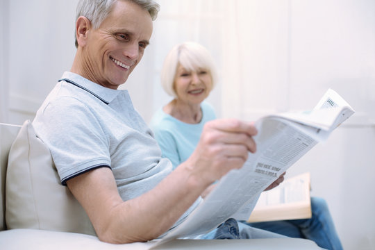 Funny article. Pleasant positive senior man laughing while sitting with his nice old wife and reading a newspaper