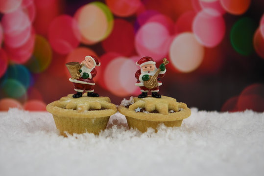 cute christmas food photography image of happy small Santa Claus decorations playing musical instruments standing on mince pies with warm red pink light pattern background and sprinkled in snow