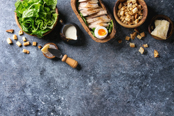 Bowls of ingredients for cooking classic Caesar salad. Sliced baked chicken breast, green roman salad, parmesan cheese, egg, croutons, salt, jug of sauce over gray texture background. Top view, space.