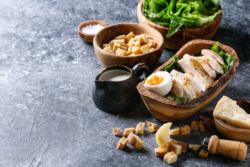 Bowls of ingredients for cooking classic Caesar salad. Sliced baked chicken breast, green roman salad, parmesan cheese, egg, croutons, salt, jug of sauce over dark gray texture background. Copy space.