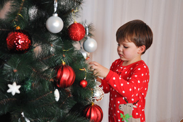 Boy in New Year's pajama decorating Christmas tree. Child putting decorations on Christmas tree