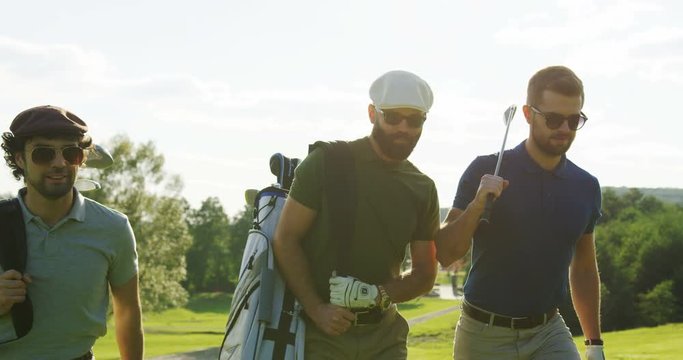 Portrait of three attractive caucasian men in sunglasses strolling with their clubs and golf bags and talking. Outdoors