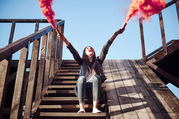 Young woman sitting on wooden steps, holding hand flares, red smoke pouring from flares, low angle view