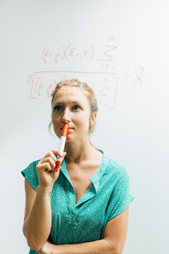 Young woman holding red marker pen gazing at complex equation on glass wall