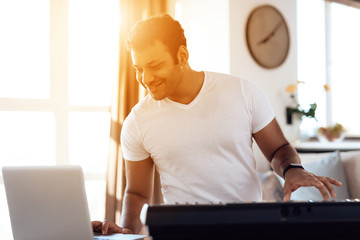 A black man sits in the living room of his apartment and plays a synthesizer. He composes music.