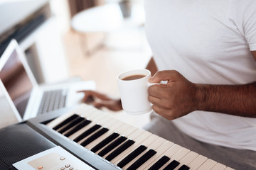A black man sits in the living room of his apartment and plays a synthesizer. He composes music and drinks coffee.