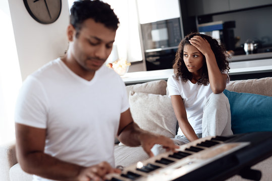 A black man sits in the living room of his apartment and plays a synthesizer. A girl sits next to him, covering her ears