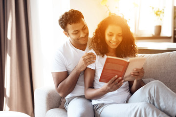 Black man and woman are sitting on the couch. A woman is reading a book, a man is sitting and hugging her.