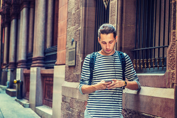 Fototapeta premium Russian Man traveling in New York, wearing striped long sleeve T shirt, shoulder carrying back bag, standing by wall on vintage street, reading, texting on cell phone, listening music with earphone..