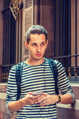 Russian Man traveling in New York, wearing striped long sleeve T shirt, shoulder carrying back bag, standing by wall on vintage street, reading, texting on cell phone, listening music with earphone..