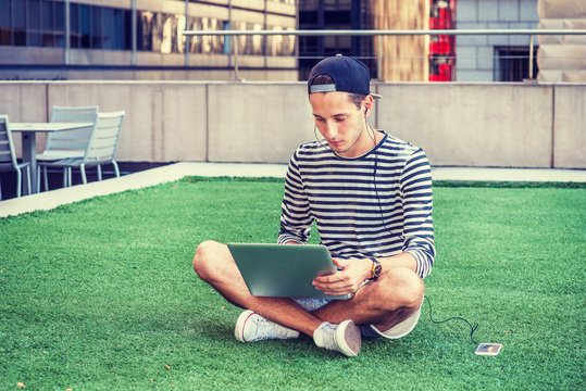 Russian man studying in New York, wearing striped long sleeve T shirt, cap backward, crossing legs, sitting on green lawn, working on laptop computer, listening music on cell phone with earphone. .