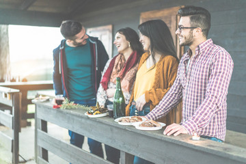 Happy friends drinking red wine and eating barbecue outside of mountain wooden house - Young people...