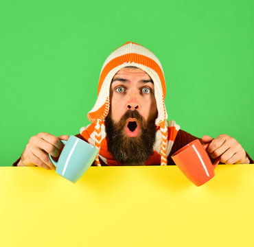 Guy with surprised face wears warm hat holding tea cups