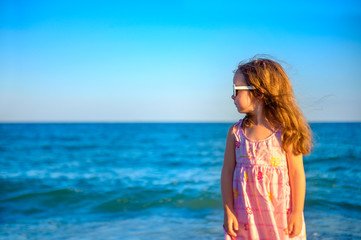 little beautiful girl in a dress standing on the beach looking away and dreaming of something.