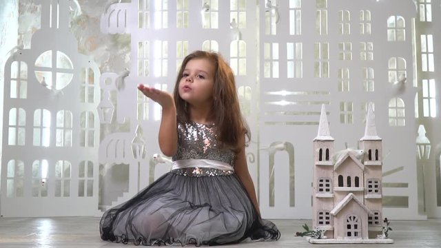 A sweet girl in a gray shiny dress sits near a toy palace and sends an air kiss, a greeting card. Slow motion shooting