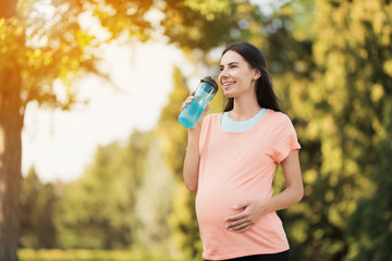 Pregnant woman walking in the park with a sports bottle in hands. She drinks from a bottle