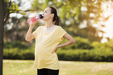 Pregnant woman walking in the park with a sports bottle in hands. She drinks from a bottle