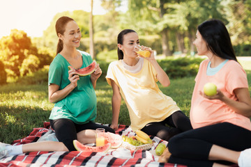 Three pregnant women sit in the park on a rug for picnics and eat. They are all smiling