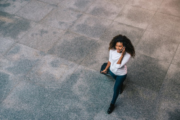 Top view portrait of young woman traveler walking on sidewalk with bag and talking on mobile phone.