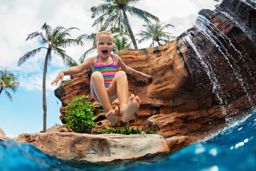 Funny child learn to swim with fun. Girl jumping high with splashes into water pool under waterfall. Healthy lifestyle, kids water sport activity, swimming lesson with parents on family beach vacation