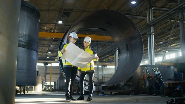 Male and Female Industrial Engineers Look at Project Blueprints While Standing Surround By Pipeline Parts in the Middle of Enormous Heavy Industry Manufacturing Factory