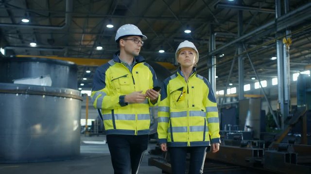 Male Project Manager with Mobile Phone and Female Industrial Engineer Having Discussion While Walking Through Heavy Industry Manufacturing Factory. 